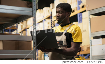 Young black employee sorting packages on shelves in a huge warehouse facility. Managing inventory with pc around packed merchandise and ready for distribution in order fulfillment. Camera B. 137747396