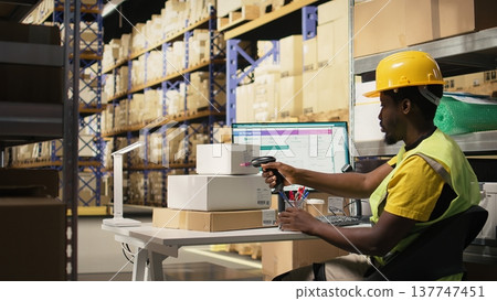 African american clerk uses barcode scanner to register shipment details tags on boxes from big industrial racks. Scanning airway bills for package tracking service, accurate logistics. Camera B. 137747451