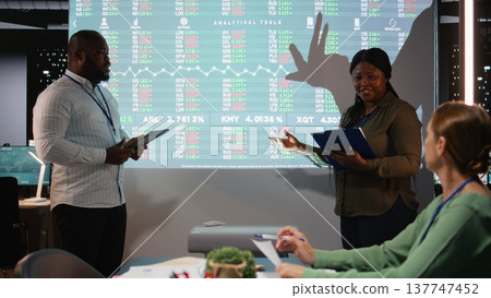 African american associates presenting their pitch to shareholders in a late night meeting, discussing about the financial situation and working for success. Evaluate supply and demand. Camera A. 137747452