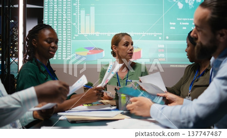 Close up of diverse staff hand over data projections papers in a meeting after hours for multinational company operations. Risk assessment and visual data to support forecasting. Camera A. Close up of diverse staff hand over data projections papers in a meeting after hours for multinational company operations. Risk assessment and visual data to support forecasting. Camera A. 137747487