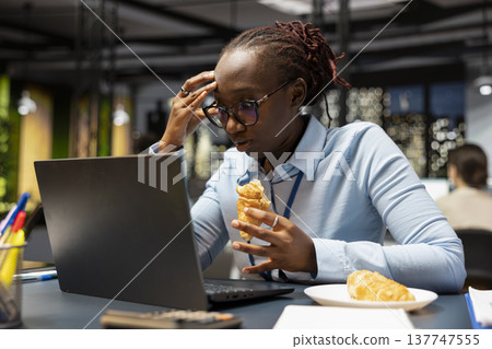 Panicked businesswoman stress eating at office desk while using laptop to finish project before deadline. Overworked African american woman snacks on croissant, doing research analysis using notebook Panicked businesswoman stress eating at office desk while using laptop to finish project before deadline. Overworked African american woman snacks on croissant, doing research analysis using notebook 137747555