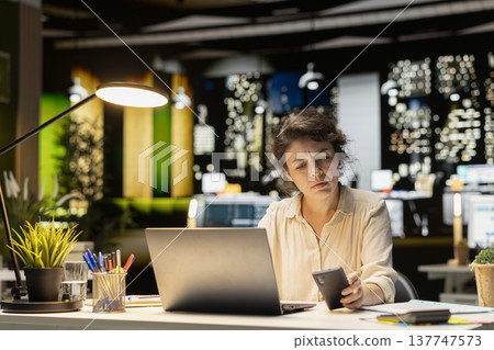 Female entrepreneur sighing and checking her messages on phone, feeling drained and frowning after being distracted by a notification in corporate office. Workplace challenges at night. 137747573