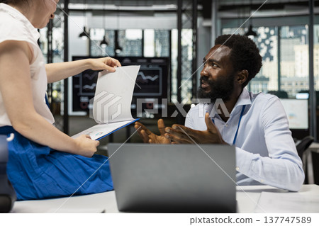 Black man and female coworker reviewing analytics, paperwork and reports at modern office desk. Multiracial business partners working on project planning, financial tasks and investment strategies. Black man and female coworker reviewing analytics, paperwork and reports at modern office desk. Multiracial business partners working on project planning, financial tasks and investment strategies. 137747589