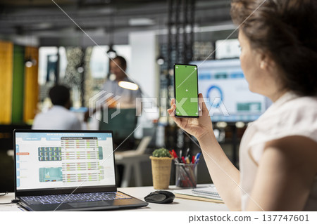 Caucasian marketing analyst using mobile device with isolated chroma key template to review business charts. Female employee viewing financial data on laptop and holding green screen smartphone. 137747601