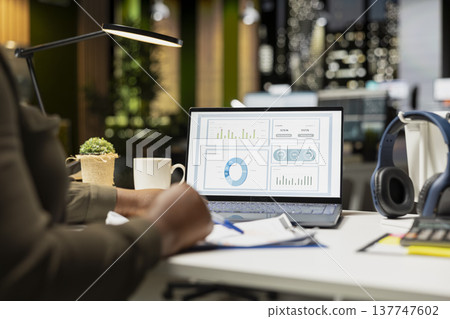 African american skilled analyst reviewing monthly reports for a new growth strategy, using infographics on laptop to plan resources allocation and align objectives with financial development. 137747602