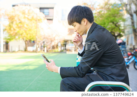一位年輕的商人坐在公園長椅上,專注地看著他的智慧型手機(上半身)。 一位年輕的商人坐在公園長椅上,專注地看著他的智慧型手機(上半身)。 137749624