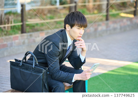 A young businessman sits on a park bench, intently looking at his smartphone (upper body). 137749625