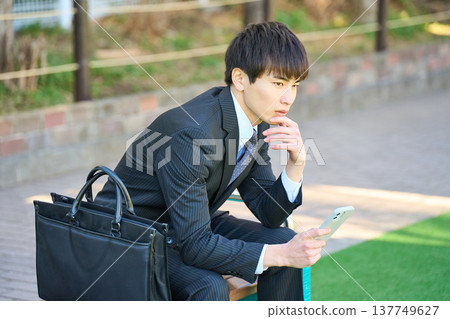A young businessman sits on a park bench, intently looking at his smartphone (upper body). A young businessman sits on a park bench, intently looking at his smartphone (upper body). 137749627