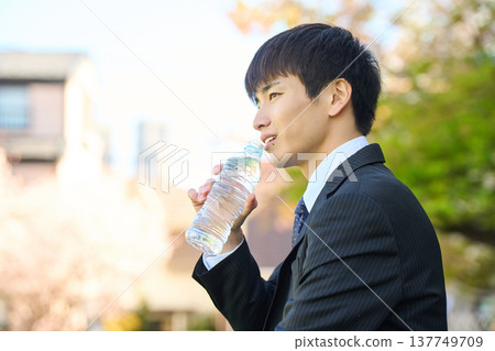 A new businessman refreshes himself by drinking bottled water in a park. (Upper body, side profile) A new businessman refreshes himself by drinking bottled water in a park. (Upper body, side profile) 137749709