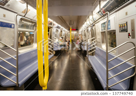 Interior view of a New York City subway train car with yellow handrails and blue seats. Public transportation scene with passengers, stainless steel doors, and urban metro commute environment. 137749740