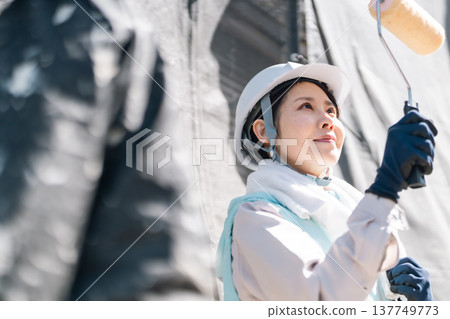 Exterior wall painting renovation. A female craftswoman, wearing a cooling vest, carefully paints the walls with a roller. Exterior wall painting renovation. A female craftswoman, wearing a cooling vest, carefully paints the walls with a roller. 137749773