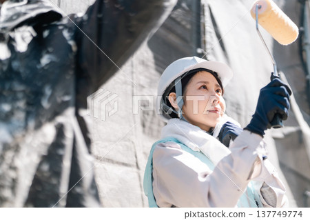 外牆粉刷翻新。一位身穿降溫背心的女工匠正用滾筒小心翼翼地粉刷牆壁。 外牆粉刷翻新。一位身穿降溫背心的女工匠正用滾筒小心翼翼地粉刷牆壁。 137749774