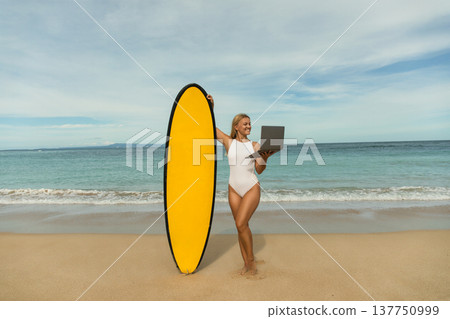 Woman enjoys digital work on beach while holding a surfboard Woman enjoys digital work on beach while holding a surfboard 137750999