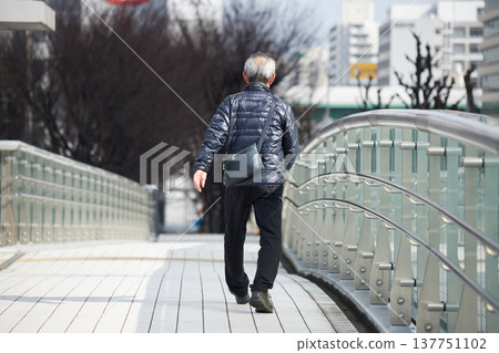 A senior man crosses a pedestrian overpass in a bustling city center on a winter day. 137751102