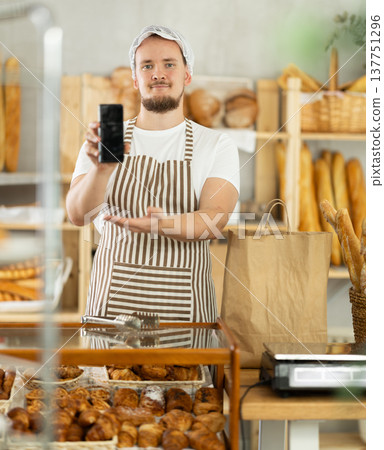 Guy seller in bakery hold out hand with phone, points at screen with finger Guy seller in bakery hold out hand with phone, points at screen with finger 137751296