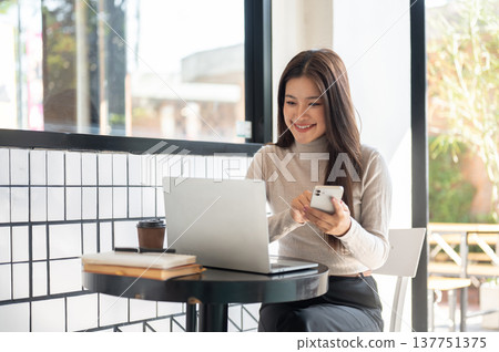 Pretty asian woman holding phone looking at laptop aside book and coffee while sits at table in cafe Pretty asian woman holding phone looking at laptop aside book and coffee while sits at table in cafe 137751375