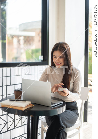 Smiling asian woman holding phone looking at laptop aside books and coffee as sits at table in cafe. Smiling asian woman holding phone looking at laptop aside books and coffee as sits at table in cafe. 137751376