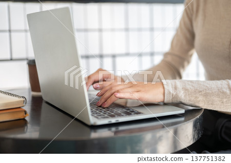 Close up of a woman's hands typing on laptop aside coffee and books while sitting at table in cafe. Close up of a woman's hands typing on laptop aside coffee and books while sitting at table in cafe. 137751382