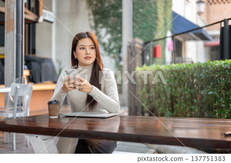 Pretty asian woman holding phone over laptop and coffee while sits at wooden table aside tree fence. Pretty asian woman holding phone over laptop and coffee while sits at wooden table aside tree fence. 137751383