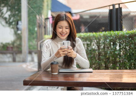 Smiling asian woman looking at phone over laptop and coffee sitting at wooden table near tree fence. Smiling asian woman looking at phone over laptop and coffee sitting at wooden table near tree fence. 137751384