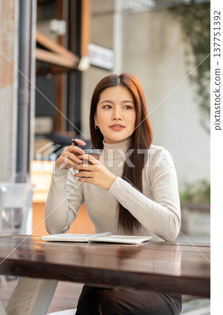 Pretty asian woman holding pen and coffee cup in both hands over book while sitting at wooden table. 137751392