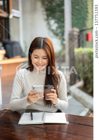 Pretty asian woman looking at phone holding coffee over notebook sits at wooden table outside cafe. Pretty asian woman looking at phone holding coffee over notebook sits at wooden table outside cafe. 137751393