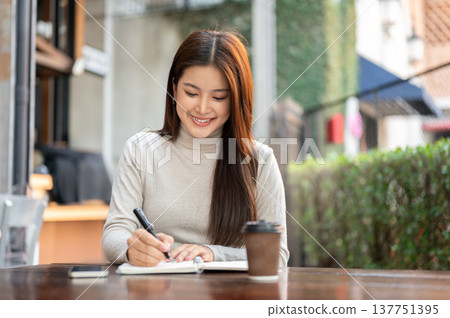 Smiling asian woman holding pen writing in notebook aside coffee sits at wooden table outside cafe. Smiling asian woman holding pen writing in notebook aside coffee sits at wooden table outside cafe. 137751395