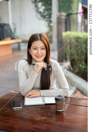 Pretty asian woman holding pen under chin thinking over book and coffee on wooden table outside cafe Pretty asian woman holding pen under chin thinking over book and coffee on wooden table outside cafe 137751396