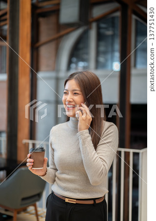 Happy asian woman in sweater holding coffee cup talking on phone while standing or walking in cafe 137751430
