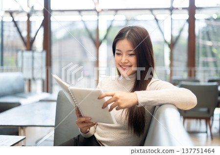 Happy asian woman holding and reading a book while sitting leaning on leather sofa or couch in cafe. Happy asian woman holding and reading a book while sitting leaning on leather sofa or couch in cafe. 137751464