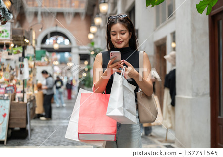 Happy asian woman carrying shopping bags looking or talking on phone while walking in mall or market Happy asian woman carrying shopping bags looking or talking on phone while walking in mall or market 137751545