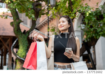Pretty asian woman holding smartphone carrying shopping bags while standing or walking in mall plaza Pretty asian woman holding smartphone carrying shopping bags while standing or walking in mall plaza 137751602