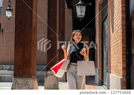 Pretty asian woman holding phone and shopping bag while walking in brick wall building with sunlight Pretty asian woman holding phone and shopping bag while walking in brick wall building with sunlight 137751608
