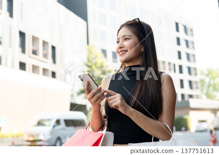 Pretty asian woman holding phone carrying shopping bags while standing or walking by street or road. Pretty asian woman holding phone carrying shopping bags while standing or walking by street or road. 137751613