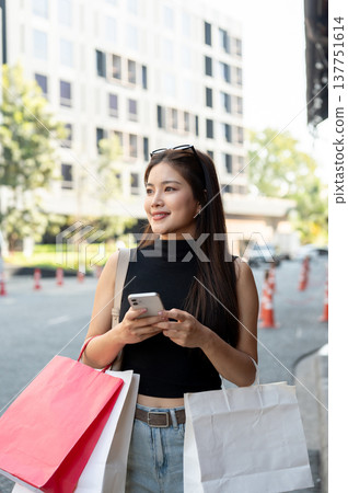 Pretty asian woman holding a phone carrying shopping bags while standing or walking by street road. 137751614