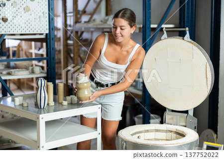 Young potter girl takes out finished ceramic dishes, various mugs and a vase from oven Young potter girl takes out finished ceramic dishes, various mugs and a vase from oven 137752022