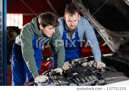 Two auto mechanics work together to diagnose an engine under the hood of a car in repair shop Two auto mechanics work together to diagnose an engine under the hood of a car in repair shop 137752135