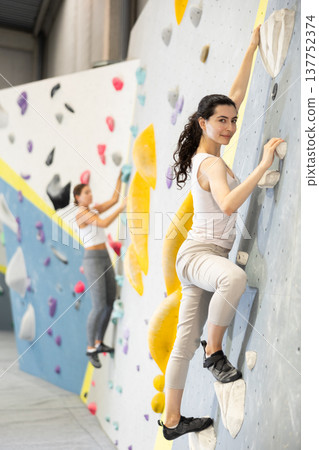 Girl is mastering climbing on training wall in gym. Woman holds on tightly to ledges and strives for top of bouldering route Girl is mastering climbing on training wall in gym. Woman holds on tightly to ledges and strives for top of bouldering route 137752374