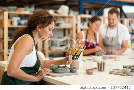 Woman learns to make designs on clay plate using brush and paint in pottery workshop Woman learns to make designs on clay plate using brush and paint in pottery workshop 137752412