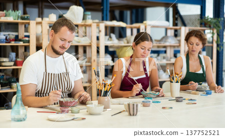 Three pottery masters work together on ceramic tableware, enjoying creative process as they paint mugs in workshop Three pottery masters work together on ceramic tableware, enjoying creative process as they paint mugs in workshop 137752521