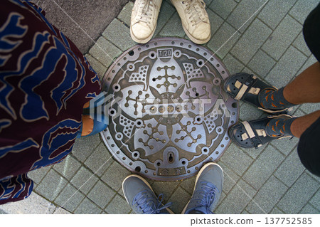 Tokyo, Japan - Oct 14 2024, close-up view of a metal manhole cover with a lotus flower depicted around it, with legs of a family of four standing around it, Ginza district, Tokyo, Japan Tokyo, Japan - Oct 14 2024, close-up view of a metal manhole cover with a lotus flower depicted around it, with legs of a family of four standing around it, Ginza district, Tokyo, Japan 137752585