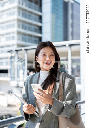 Pretty asian woman student office worker holding tablet and phone walking on bridge with cityscape. 137753063