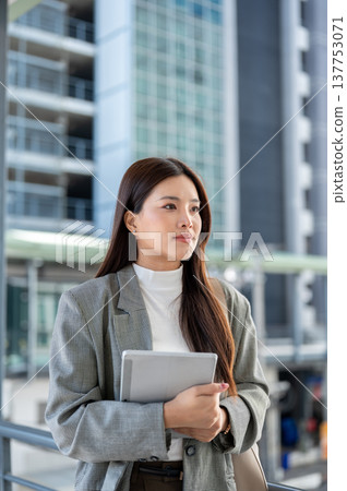 Asian woman student office worker holding or carrying tablet standing on footbridge with cityscape. Asian woman student office worker holding or carrying tablet standing on footbridge with cityscape. 137753071