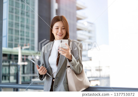 Asian woman student office worker holding tablet looking at phone standing on bridge with cityscape. 137753072