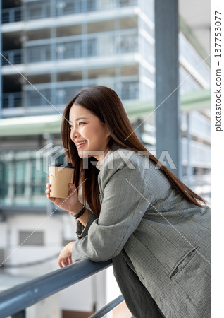 Asian woman office worker holding cup drinking coffee leaning over railing of bridge with cityscape. Asian woman office worker holding cup drinking coffee leaning over railing of bridge with cityscape. 137753077