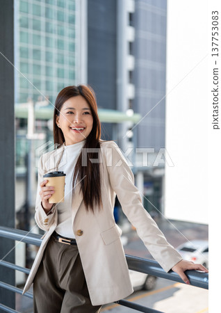 Smiling asian woman office worker holding coffee cup standing aside railing on bridge with cityscape 137753083