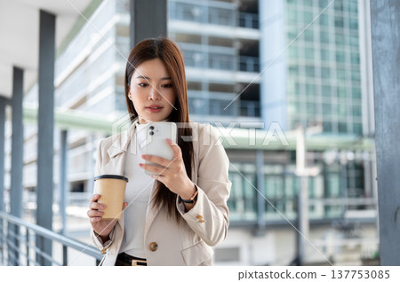 Asian woman office worker holding coffee cup looking at phone standing on footbridge with cityscape. 137753085