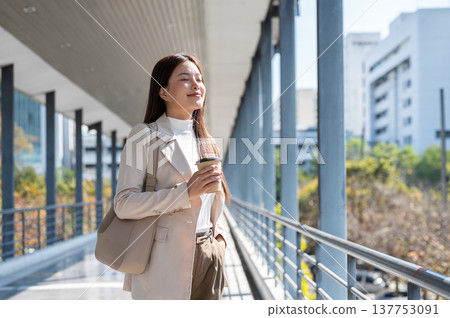 Pretty asian woman office worker closing eyes holding coffee and standing in sunlight on footbridge. 137753091