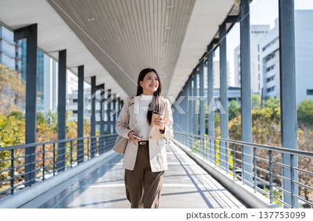 Smiling asian woman office worker holding coffee and phone looking around as walking on footbridge. 137753099