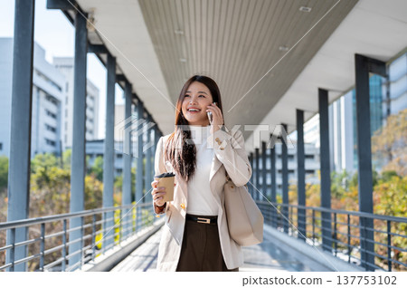 Pretty asian woman office worker holding coffee talking on phone as walking standing on a footbridge Pretty asian woman office worker holding coffee talking on phone as walking standing on a footbridge 137753102